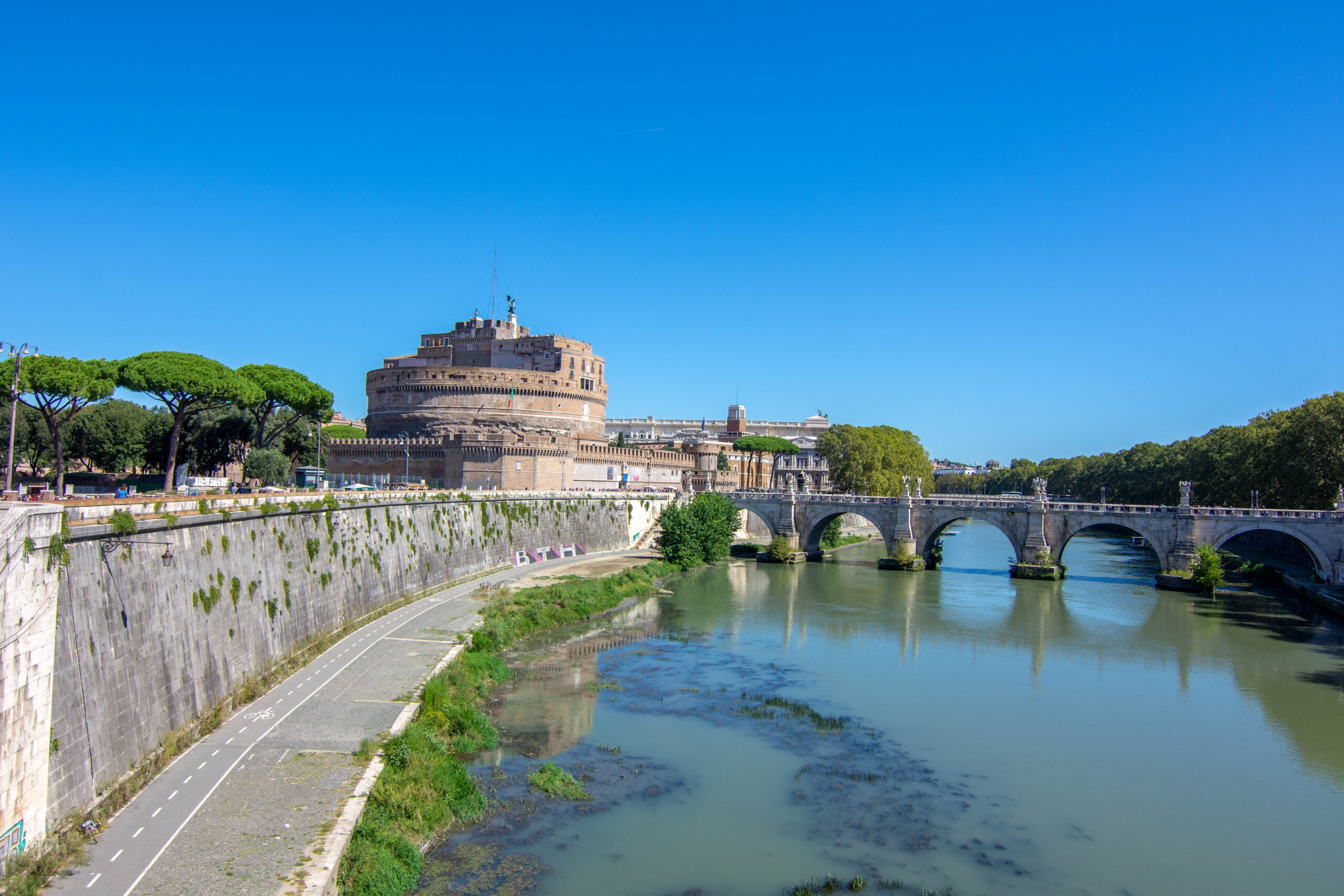 Castel Sant'Angelo beside the Tiber River with a stone bridge, green trees, and a clear blue sky above.