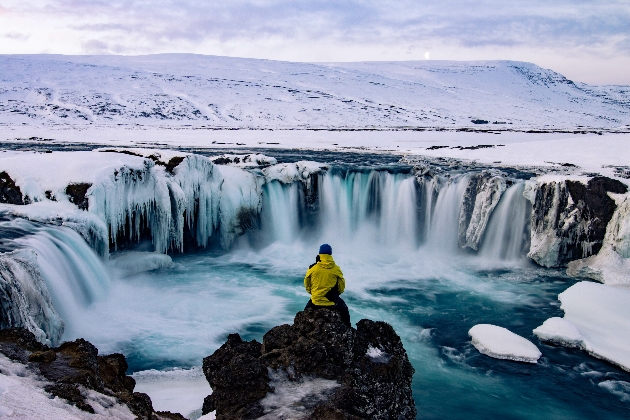 Person in a bright yellow jacket sits on rocks beside a frozen waterfall in a snowy landscape.